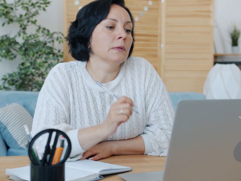 Žena srednjih godina sedi za laptopom, za ra radnim stolom na kom je kancelarijski pribor i notes i razmišlja