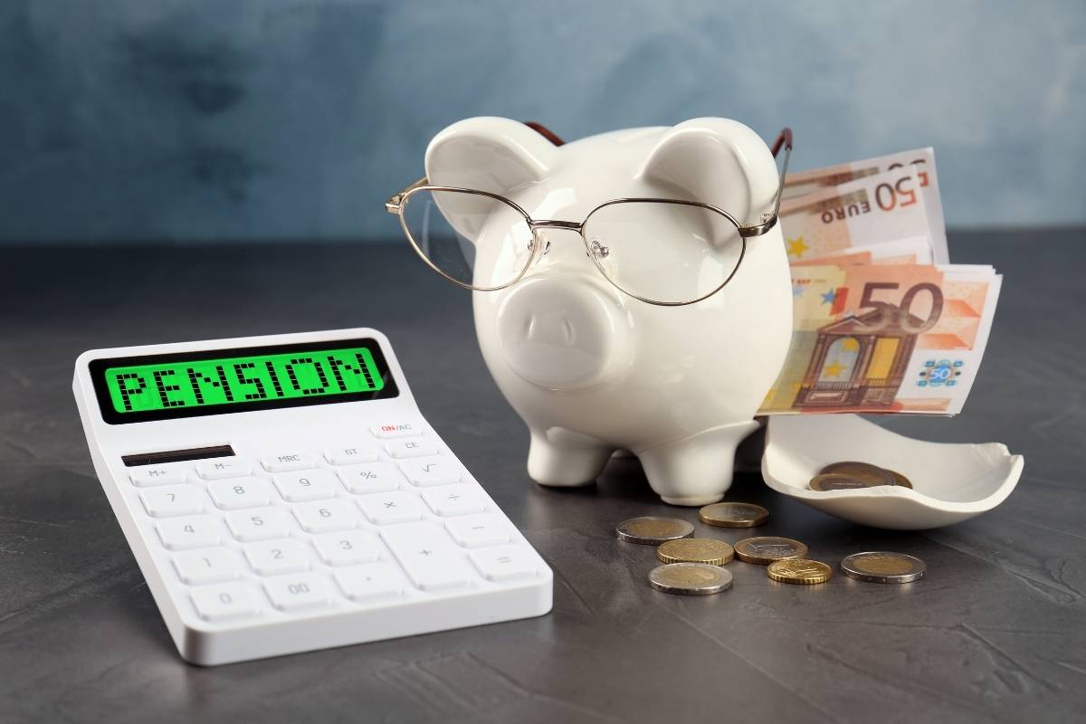 Kasica-prasica sa naočarima stoji pored kalkulatora koji prikazuje „PENSION”, uz evre i kovanice na stolu – simbol štednje i planiranja penzije.